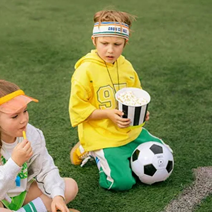 Two children on a grassy field, one holding popcorn and the other eating, with a soccer ball nearby.