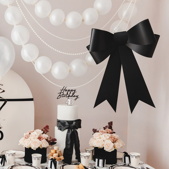 A large black bow decoration placed on a table with a birthday cake and floral arrangements, with hanging white balloons in the background.