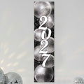 A vertical rectangular wall art print featuring the year '2027' in large white script over a black background with mirrored disco ball illustrations. The print is shown hanging against a light gray wall with decorative disco balls at the bottom corners.