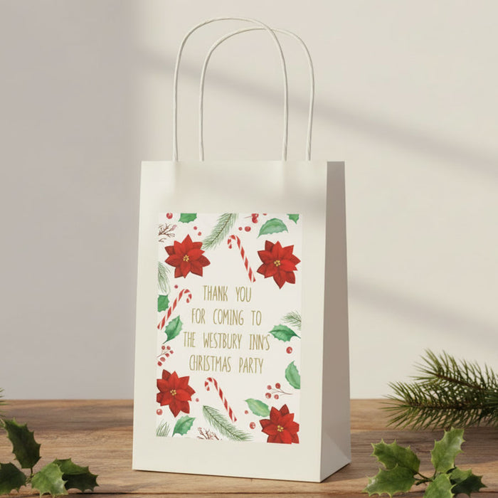 A white paper gift bag with white twisted handles stands on a wooden surface against a light beige wall. The front of the bag has a festive printed design with red poinsettias, green leaves, candy canes, and gold text reading, 'THANK YOU FOR COMING TO THE WESTBURY INNS CHRISTMAS PARTY.