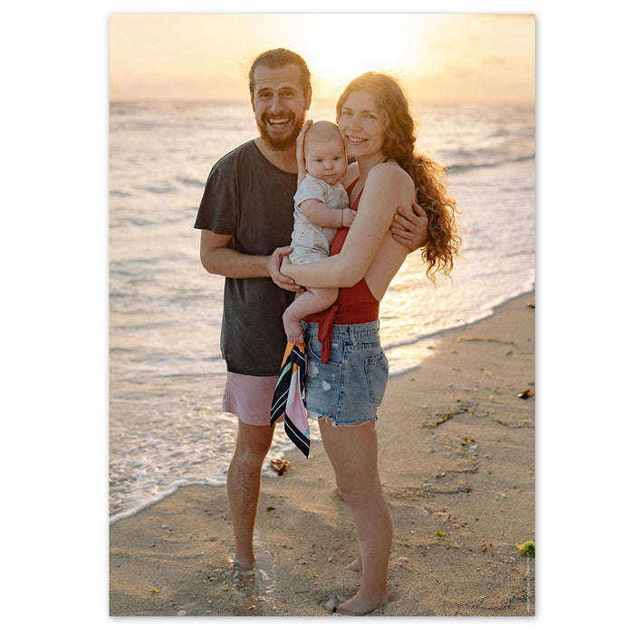 A family of three, two adults and a baby, smiling and standing on a beach at sunset. The adults are holding the baby, and they appear to be enjoying a happy moment together.